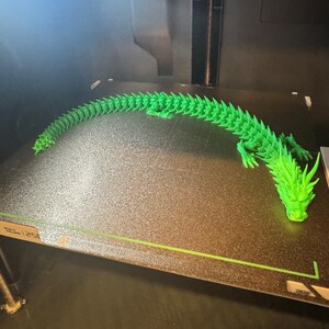 May include: A vibrant green, 3D-printed dragon, articulated for movement. The dragon is positioned on a dark surface, with its head and tail in view. The image is taken inside a 3D printer, showcasing the model's detail.