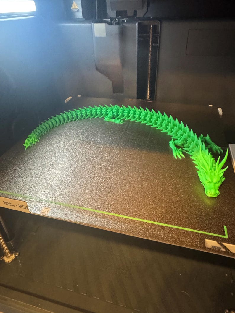 May include: A vibrant green, 3D-printed dragon, articulated for movement. The dragon is positioned on a dark surface, with its head and tail in view. The image is taken inside a 3D printer, showcasing the model's detail.