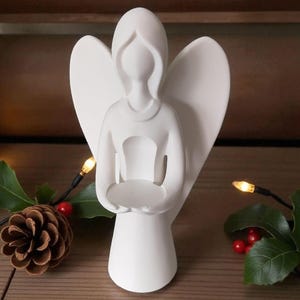 May include: A white ceramic angel figurine with outstretched wings, holding a small, round dish. The angel has a simple, elegant design. A pinecone, holly, and string lights are in the background, suggesting a holiday theme.