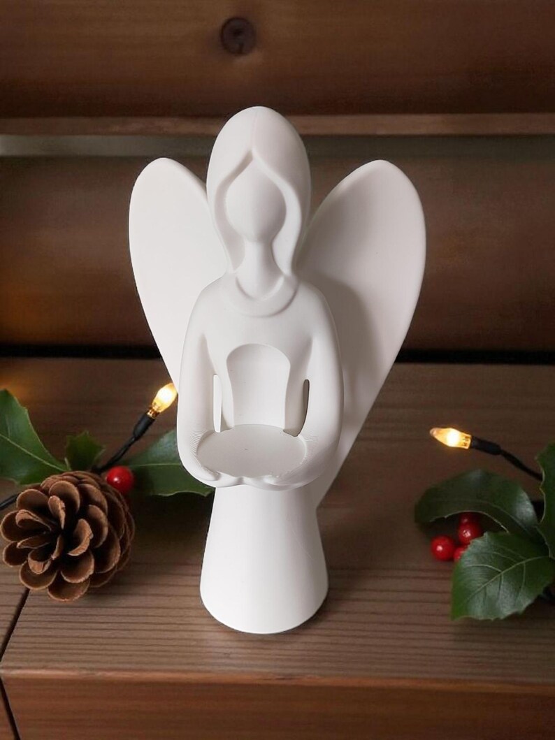 May include: A white ceramic angel figurine with outstretched wings, holding a small, round dish. The angel has a simple, elegant design. A pinecone, holly, and string lights are in the background, suggesting a holiday theme.