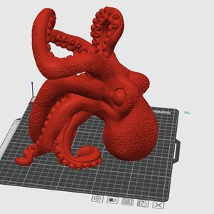 May include: A red octopus figurine with detailed tentacles and a textured surface. The octopus is positioned on a black and gray grid, likely a 3D printing bed. The figurine is a solid, three-dimensional object.