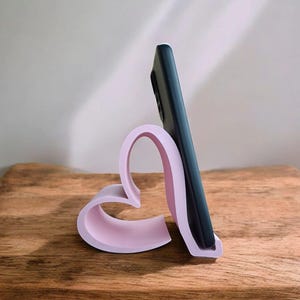 May include: A pink heart-shaped phone stand holds a black smartphone in a vertical position. The stand is on a wooden surface.