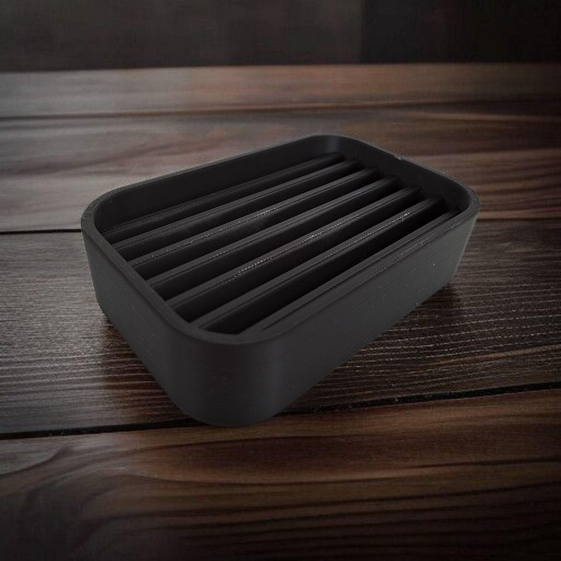 May include: A rectangular, black soap dish with a slatted design. The dish is made of a matte material and sits on a wooden surface. The soap dish is designed to allow water to drain, extending the life of the soap.
