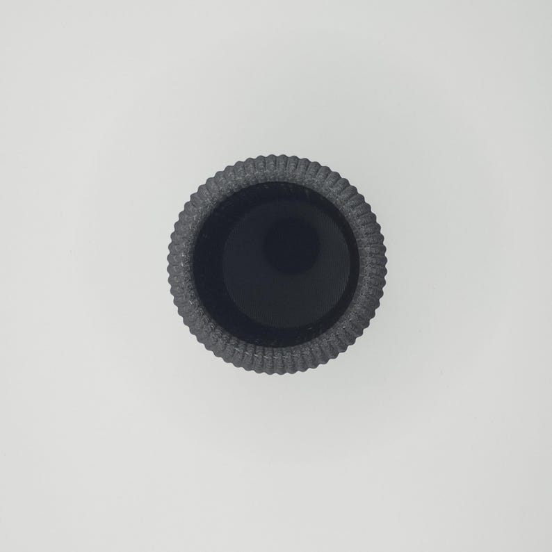 May include: A top-down view of a dark gray, textured, round container. The container has a ribbed design around the exterior and a deep, black interior. The container is centered on a white background.