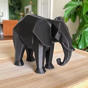 Op de afbeelding: Een zwarte geometrische olifant sculptuur met een polygonale vormgeving. De olifant staat op een houten oppervlak. De sculptuur heeft een gedetailleerde slurf, oren en poten. De achtergrond omvat een groene plant en een glimp van een kamer.
