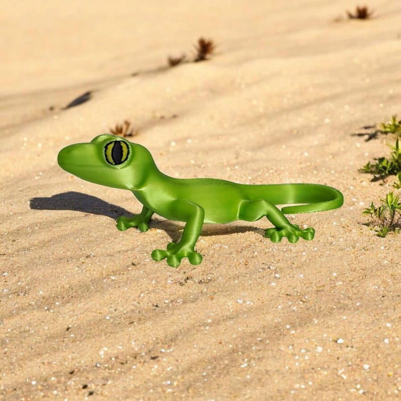 May include: A bright green gecko figurine with a detailed design, including visible toes and a curled tail. The gecko has large, black and yellow eyes. It is positioned on a sandy surface, with small plants in the background.