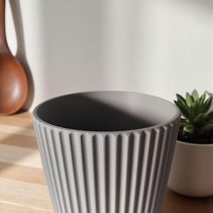 May include: A gray, ribbed ceramic planter with a wide opening. The planter has vertical grooves and a slightly textured surface. A small white pot with a green succulent is to the right.