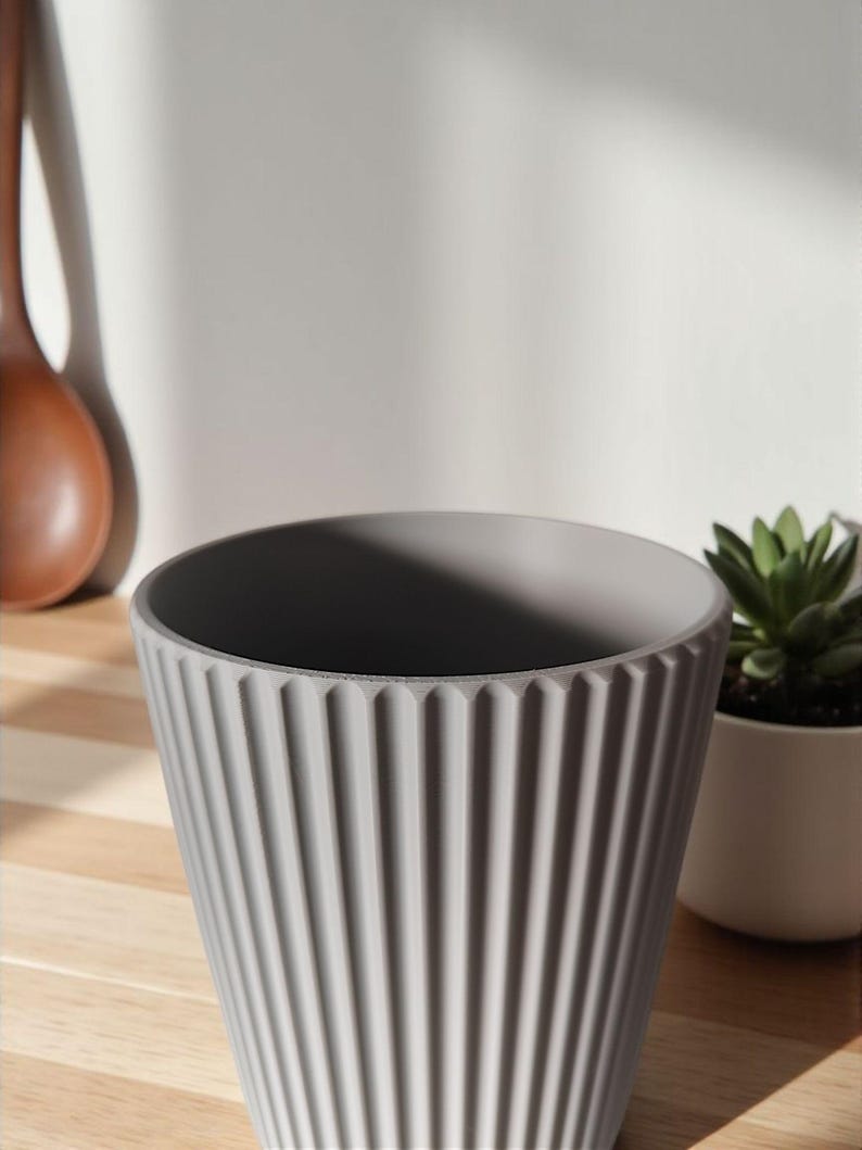 May include: A gray, ribbed ceramic planter with a wide opening. The planter has vertical grooves and a slightly textured surface. A small white pot with a green succulent is to the right.