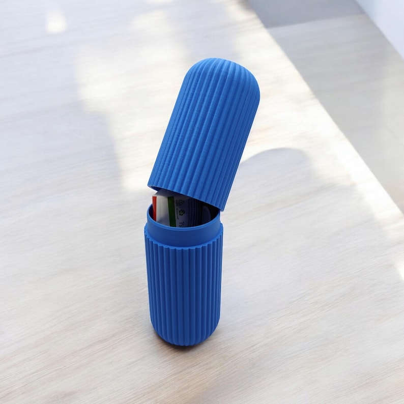 May include: A blue, ribbed toothbrush travel case with the lid slightly ajar. The cylindrical case is made of a solid blue material and sits on a light-colored wooden surface. The lid is open, revealing the contents inside.