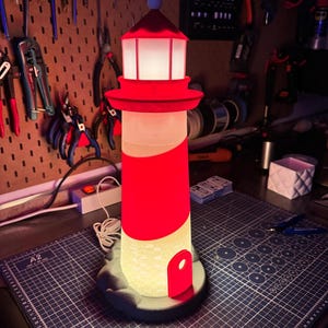 May include: A red and white lighthouse-shaped lamp with a glowing interior. The lamp has a red top and a red stripe around the body. The base is dark gray. The lamp is on a gray cutting mat.
