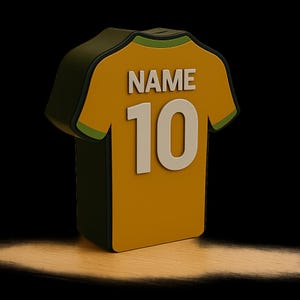 K&ouml;nnte beinhalten: 3D-Darstellung eines gelb-gr&uuml;nen Fu&szlig;balltrikots mit dem Wort "NAME" &uuml;ber der wei&szlig;en Nummer "10". Das Trikot hat eine dunkelgr&uuml;ne Kontur und ist vor einem schwarzen Hintergrund mit einem Spotlight-Effekt platziert.