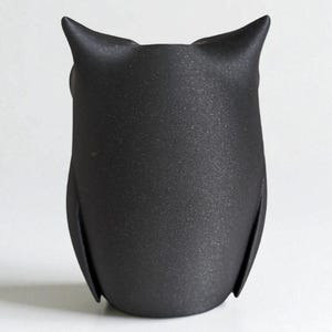 Op de afbeelding: Een zwarte uilfiguur met een getextureerd oppervlak. De uil heeft puntige oren en een afgerond lichaam. De figuur staat rechtop tegen een witte achtergrond.