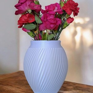 Puede incluir: Un jarr&oacute;n azul claro con un dise&ntilde;o acanalado en espiral, que contiene un ramo de flores rojas y magenta. Las flores tienen tallos y hojas verdes. El jarr&oacute;n est&aacute; sobre una superficie de madera.