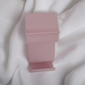 May include: A light pink, rectangular phone stand with a tiered design. The stand has a small ledge at the bottom and a horizontal ridge near the top. The stand is made of a solid material and sits on a white, textured surface.