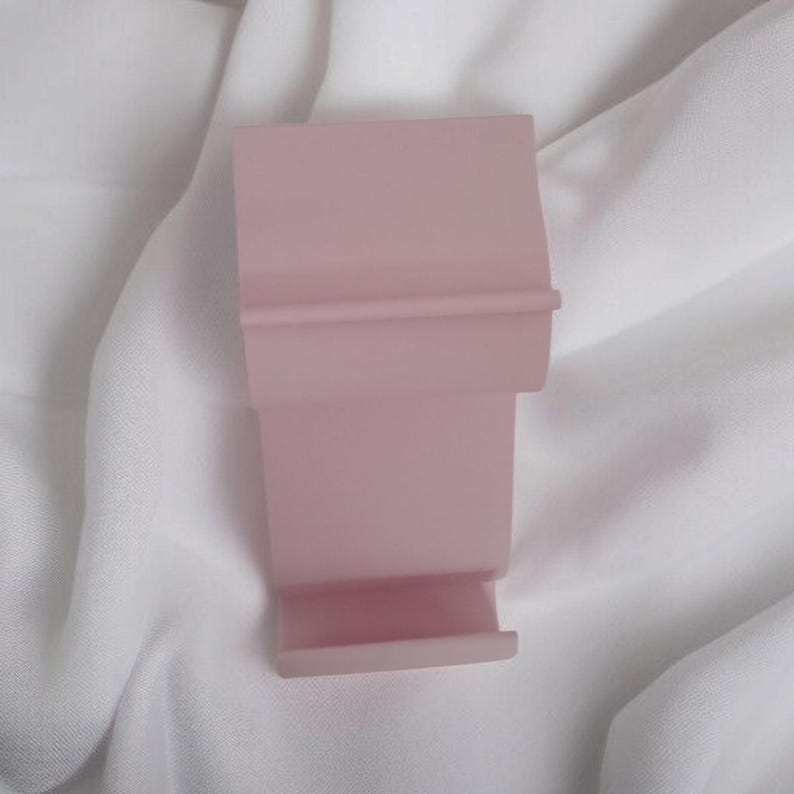 May include: A light pink, rectangular phone stand with a tiered design. The stand has a small ledge at the bottom and a horizontal ridge near the top. The stand is made of a solid material and sits on a white, textured surface.