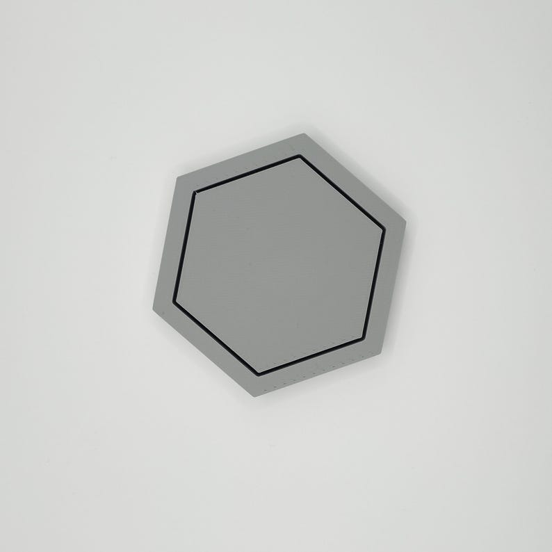 May include: A gray, geometric coaster with a black border. The coaster has a hexagonal shape and is set against a white background. The coaster is a solid color and has a simple, modern design.