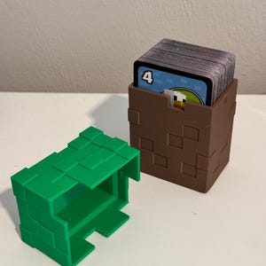 May include: Two 3D-printed card holders. One is green and open, the other is brown and holds a deck of cards. The brown holder has a pixelated design. The cards have a blue border and the number 4.