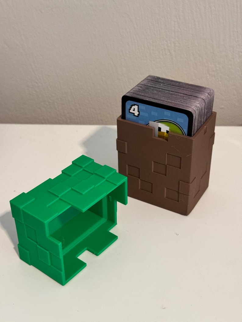 May include: Two 3D-printed card holders. One is green and open, the other is brown and holds a deck of cards. The brown holder has a pixelated design. The cards have a blue border and the number 4.