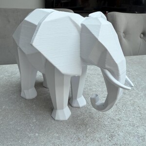 Op de afbeelding: Een witte, geometrische olifant sculptuur met een polygonale vormgeving. De olifant heeft een slurf, slagtanden en vier poten. De sculptuur is gemaakt van een solide, ondoorzichtig materiaal en staat op een grijze ondergrond.