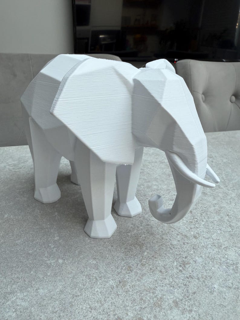 Op de afbeelding: Een witte, geometrische olifant sculptuur met een polygonale vormgeving. De olifant heeft een slurf, slagtanden en vier poten. De sculptuur is gemaakt van een solide, ondoorzichtig materiaal en staat op een grijze ondergrond.