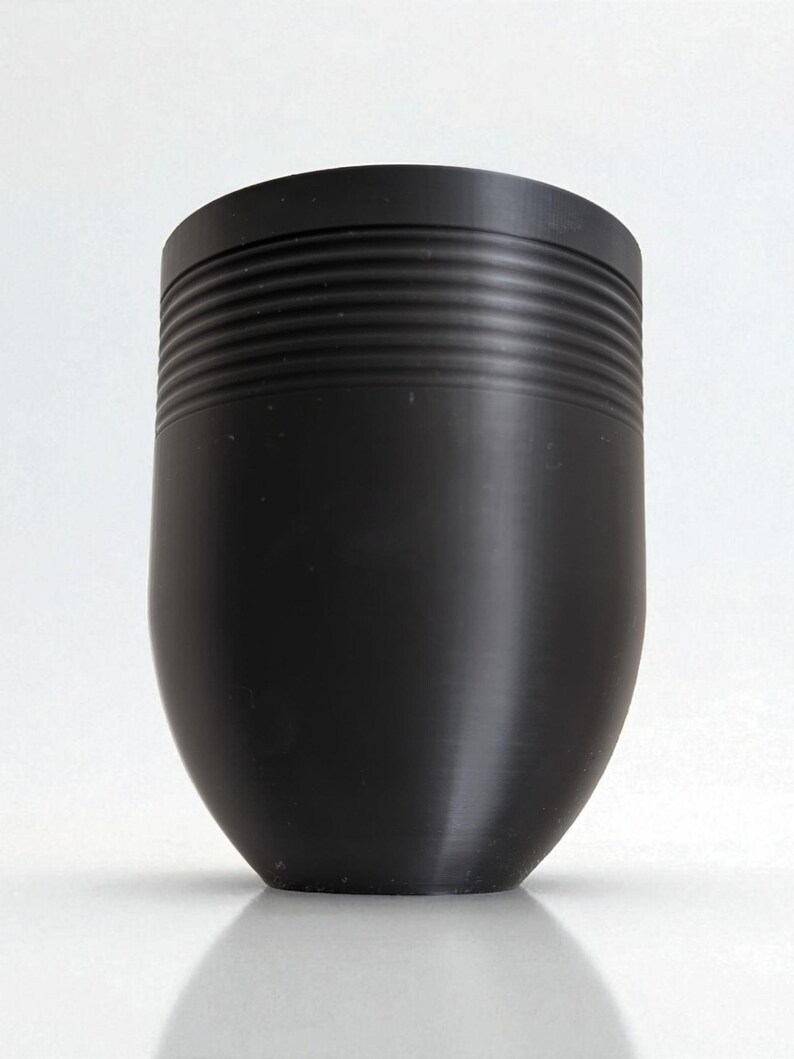 May include: A black, cylindrical object with a rounded base. The upper portion features a series of horizontal, ridged lines. The object has a smooth, matte finish and is set against a white background.