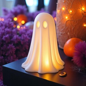 May include: A glowing white ghost-shaped lamp with two oval eye cutouts. The lamp is on a black surface, with purple flowers and an orange pumpkin in the background. A small gold-colored charm is in the foreground.