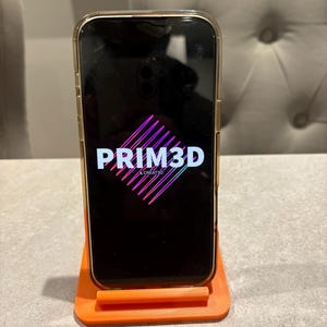 May include: A smartphone in a gold case is displayed on an orange phone stand. The phone screen displays the text "PRIM3D & CREATE3D" in white and pink. The stand is rectangular with a lip to hold the phone.