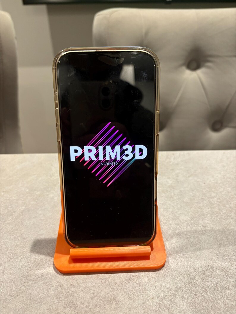May include: A smartphone in a gold case is displayed on an orange phone stand. The phone screen displays the text "PRIM3D & CREATE3D" in white and pink. The stand is rectangular with a lip to hold the phone.
