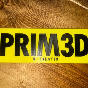 K&ouml;nnte beinhalten: Ein rechteckiges gelbes Schild mit dem Text "PRIM3D" in gro&szlig;en, schwarzen 3D-Buchstaben. Darunter steht in kleinerer Schrift "& CREATE3D". Das Schild liegt auf einer Holzoberfl&auml;che.