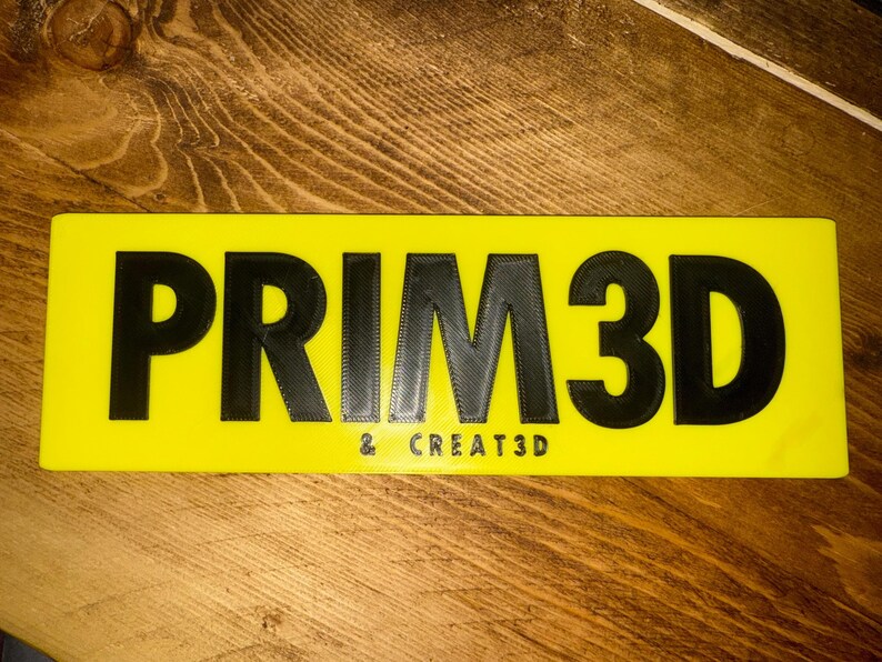 K&ouml;nnte beinhalten: Ein rechteckiges gelbes Schild mit dem Text "PRIM3D" in gro&szlig;en, schwarzen 3D-Buchstaben. Darunter steht in kleinerer Schrift "& CREATE3D". Das Schild liegt auf einer Holzoberfl&auml;che.
