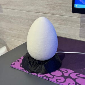 May include: A white, egg-shaped decorative lamp sits on a black, textured base. The lamp is on a purple and black patterned mousepad. The lamp is plugged into a computer.