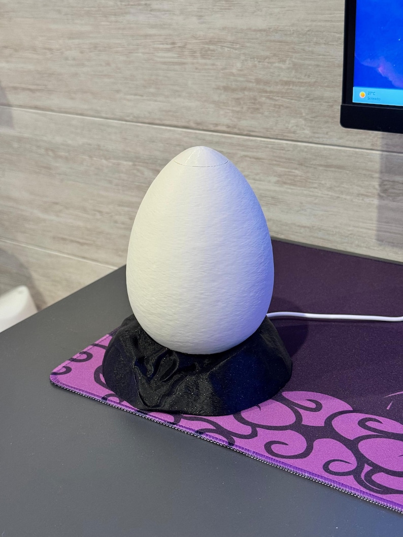 May include: A white, egg-shaped decorative lamp sits on a black, textured base. The lamp is on a purple and black patterned mousepad. The lamp is plugged into a computer.