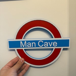 May include: A sign with a red circle and a blue rectangular bar, resembling a subway logo, with the words "Man Cave" in white. The sign is held against a white wall.