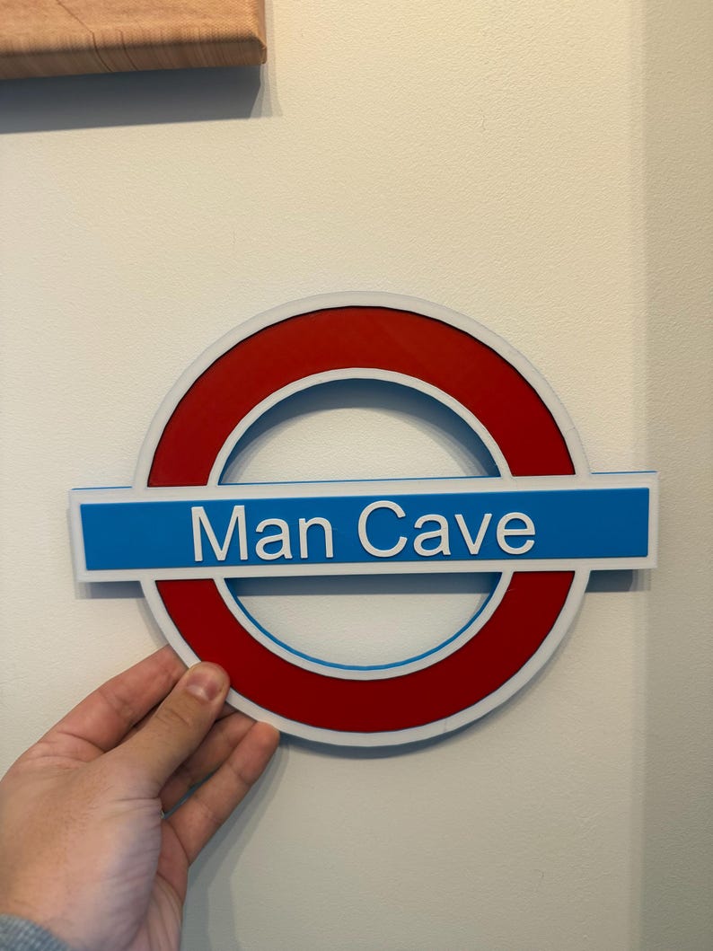 May include: A sign with a red circle and a blue rectangular bar, resembling a subway logo, with the words "Man Cave" in white. The sign is held against a white wall.