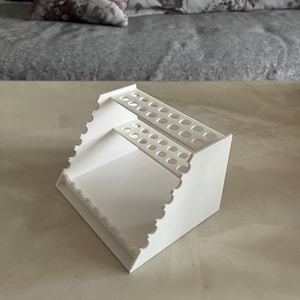 May include: A white, tiered organizer with multiple circular holes for storage. The organizer has a scalloped edge and is designed to hold various items. The design is simple and functional, suitable for a desk or craft area.