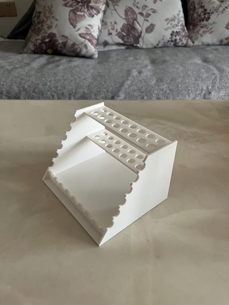 May include: A white, tiered organizer with multiple circular holes for storage. The organizer has a scalloped edge and is designed to hold various items. The design is simple and functional, suitable for a desk or craft area.