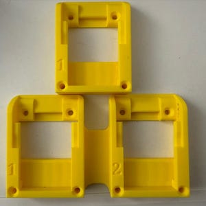 May include: Three bright yellow plastic rectangular frames with rounded corners. Each frame has a central opening and several small holes. Numbers 1 and 2 are printed on the frames.