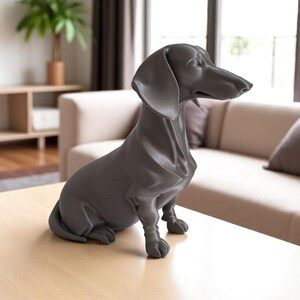 May include: A dark gray dachshund dog statue sitting on a light wood table. The dog is in a seated position with its head turned slightly to the right. The statue has a smooth finish and is a decorative item.