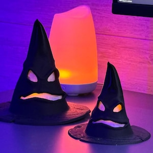 Op de afbeelding: Twee zwarte, Halloween-thema sorteerhoeden met uitgesneden ogen en monden. Een oranje en witte diffuser op de achtergrond straalt een warme gloed uit. Op een paars oppervlak.