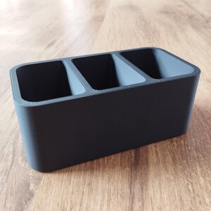 May include: A rectangular, matte black organizer with three equally sized compartments. The interior of each compartment is a lighter shade of gray. The organizer is made of a solid material and sits on a wooden surface.