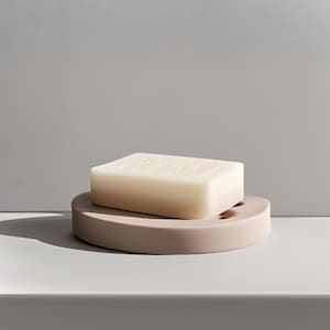 May include: A rectangular bar of light-colored soap rests on a round, taupe-colored soap dish. The soap dish has a curved design with small drainage holes. The background is a solid, neutral gray.