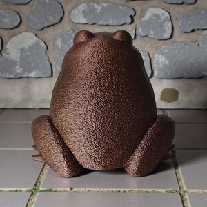 May include: A bronze-colored frog statue with a textured surface, viewed from the back. The frog is in a seated position with its legs spread. The background features a stone wall.