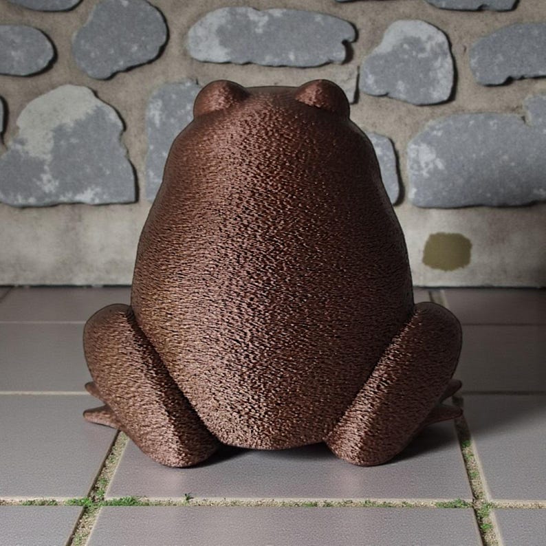 May include: A bronze-colored frog statue with a textured surface, viewed from the back. The frog is in a seated position with its legs spread. The background features a stone wall.