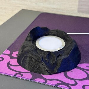 May include: A black, volcano-shaped wireless charger with a silver ring around the charging surface. The charger sits on a purple and black patterned mousepad. A white charging cable extends from the charger.