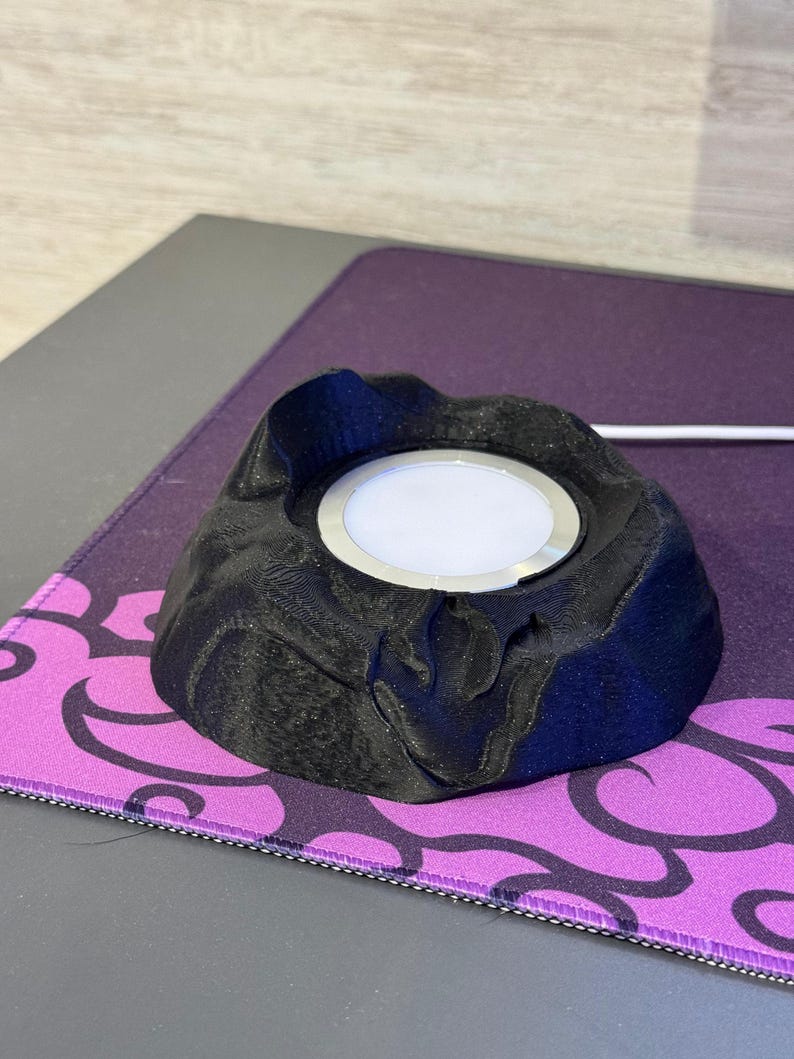May include: A black, volcano-shaped wireless charger with a silver ring around the charging surface. The charger sits on a purple and black patterned mousepad. A white charging cable extends from the charger.