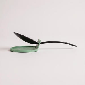 May include: A modern kitchen spoon rest in a minimalist design. The spoon rest features a round, light green base with a black spoon and handle. The spoon is angled upwards, and the handle curves gracefully.
