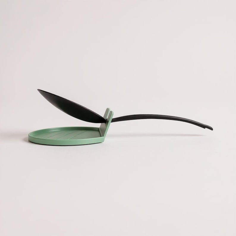 May include: A modern kitchen spoon rest in a minimalist design. The spoon rest features a round, light green base with a black spoon and handle. The spoon is angled upwards, and the handle curves gracefully.
