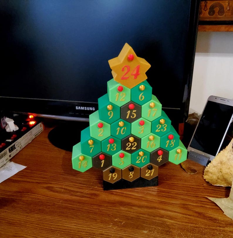 May include: A green and brown Christmas tree-shaped advent calendar with a gold star on top, displaying the number 24. The calendar has hexagonal compartments numbered 1-24, with red and gold accents. The base is black.