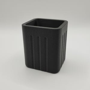 May include: A textured, matte black square container with vertical grooves on the sides. The open top reveals a hollow interior. The container is designed with a minimalist aesthetic.