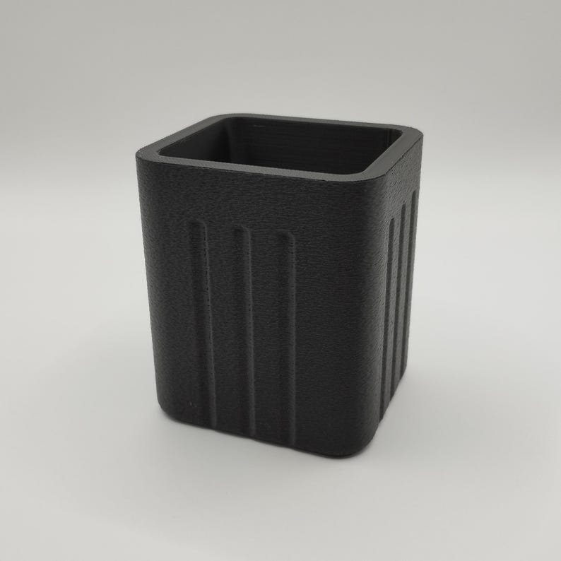May include: A textured, matte black square container with vertical grooves on the sides. The open top reveals a hollow interior. The container is designed with a minimalist aesthetic.